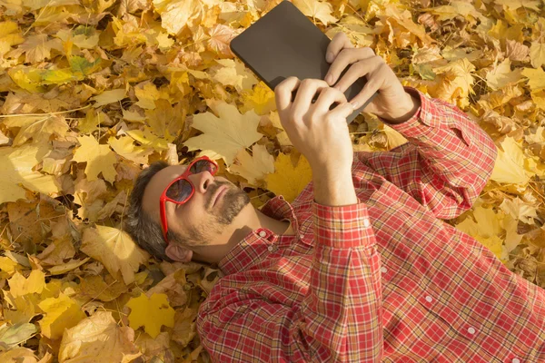 Man lying on the ground covered with yellow leaves and using digital tablet device