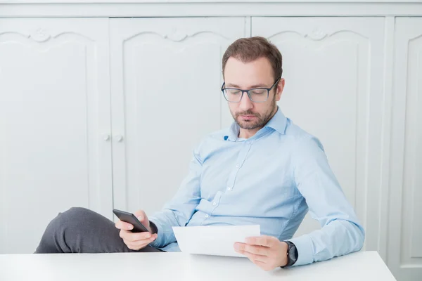Businessman sitting at his desk, looking at paper document and holding smart phone