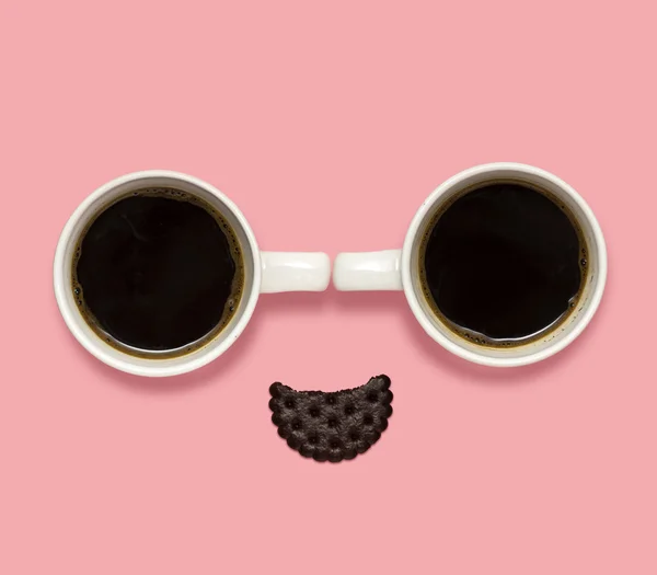 Coffee smile. Creative concept photo of coffee cups with a cracker in the shape of a smiling face on pink background.