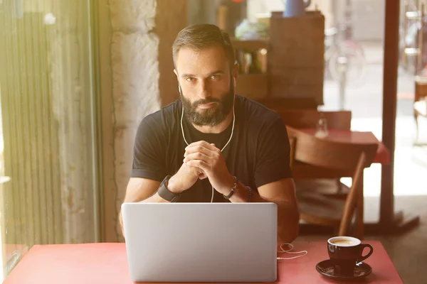 Young Bearded Businessman Wearing Black Tshirt Working Laptop Urban Cafe.Man Sitting Wood Table Cup Coffee Listening Music.Coworking Process Business Startup.Blurred Background.Sunlight effect.