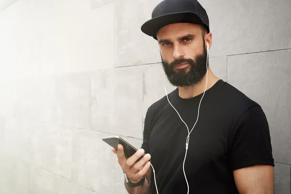 Bearded Muscular Man Wearing Black Tshirt Blank Snapback Cap Summer Time.Young Men Smiling Opposite Empty Gray Concrete Wall Background Using Smartphone Headphones.Horizontal Mockup.Color filter.