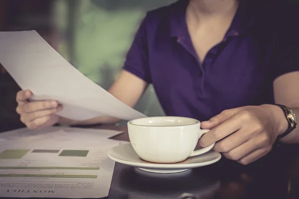 Close up hand of business women holding cup of coffee and paper graph chart  on wooden desk indoor office. vintage filter effect.