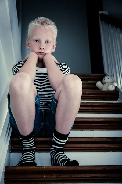 Sad blond boy sits on stairs with elbows on knees