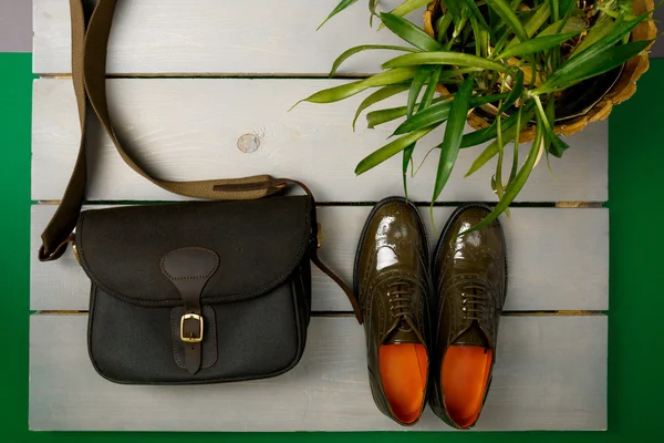 Green lacquered oxford shoes and crossbody bag on wooden background near flower pot. Top view. Close up. Copy space