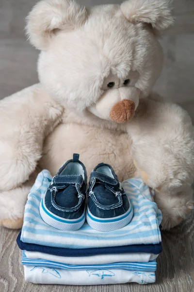 Folded blue and white bodysuit with shoes on it near big teddy bear  grey wooden background. diaper for newborn boy. Stack of infant clothing. Child outfit. Copy space.