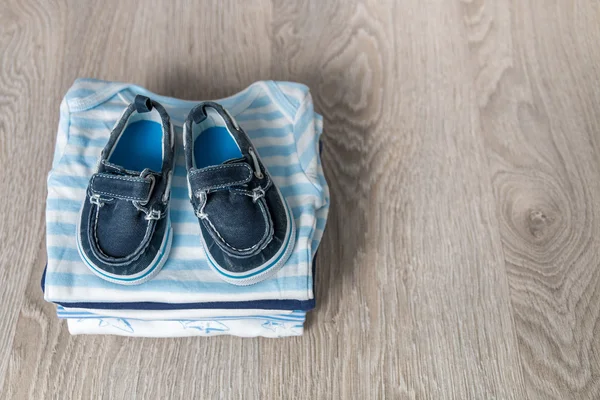 Folded blue and white bodysuit with shoes on it  grey wooden background. diaper for newborn boy. Stack of infant clothing. Child outfit. Copy space.