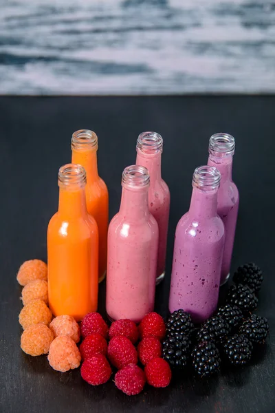 Six bottles with smoothies and raspberry, red, yellow, blackberry on black table grey background. Milk shake in glass jar berries. diet or vegan food concept, fresh vitamin
