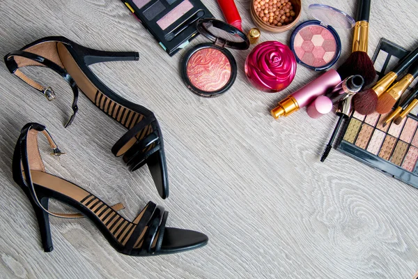 Set of decorative cosmetics and brushes near black heeled sandals on grey wooden background. Various makeup products. Top view, frame. Copy space.