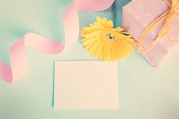 Pink dotted gift box, yellow gerbera flower and empty card over a blue background. Vintage effect filter.