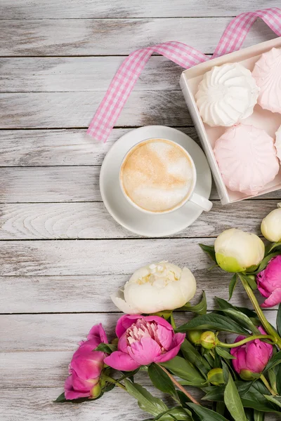 A cup of coffee next to bouquet of peonies, a box with marshmallows and pink ribbon on wooden background. Top view.