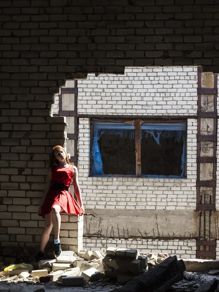 Red-haired thin slim girl with freckles  in red dress on  background of  wall of  destroyed building