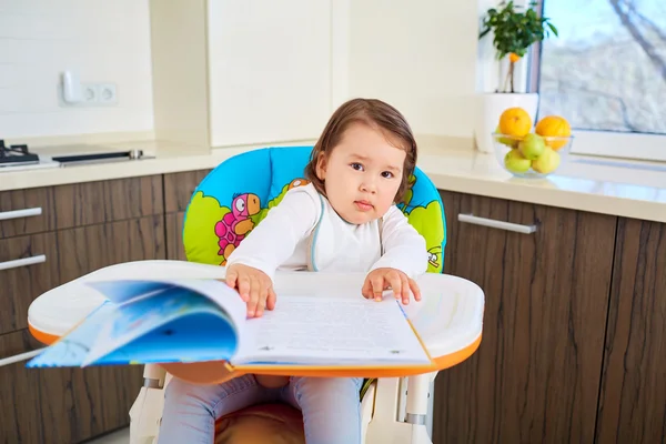 Funny toddler girl reading a book in kitchen