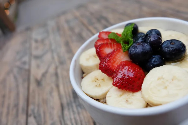 Acai bowl with fresh fruit strawberry, blueberry, banana and peppermint leaves on top on the wooden table.