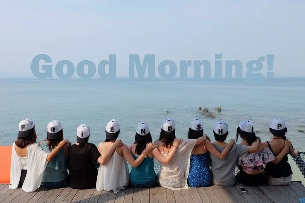 Group of ten women friends sit hug together look Good Morning! blue sea sky.  They wear same design caps with FRIENDSHIP alphabets.