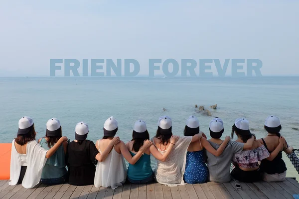 Group of ten women friends sit hug together look FRIEND  FOREVER blue sea sky. They wear same design black and white caps.