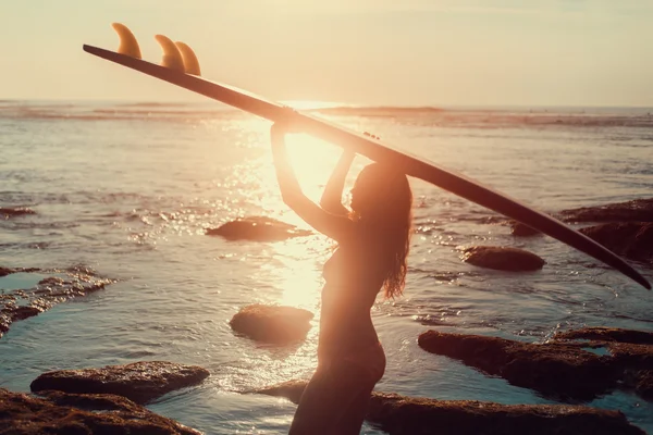 Silhouette surfer woman in bikini on tropical beach holding surfboard at sunset