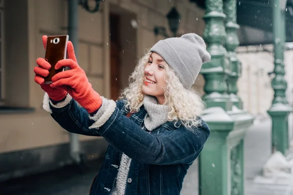 Young curly blond girl in red gloves, grey hat and winter jeans jacket is taking a selfie in the street