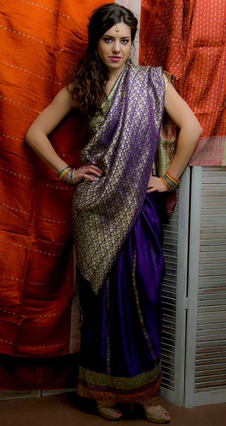 The young dark-haired woman in the rich Indian saris standing akimbo on the background screen decorated with bright silk. Indian style.
