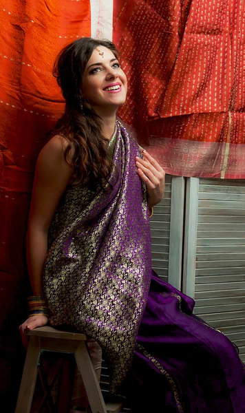 The young dark-haired woman in a beautiful Indian saris laughing happily sitting on a background of bright draped screen. Indian style.