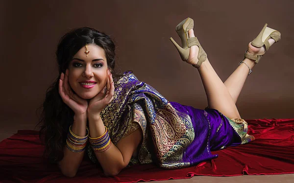 The young dark-haired woman in the rich Indian sari lying on bright red velvet and smiles placing hands under the face. Indian style.