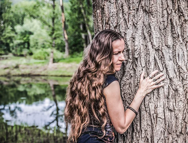 Young woman with long golden hair thoughtfully pressed to the trunk of a huge tree on the background of forest lake