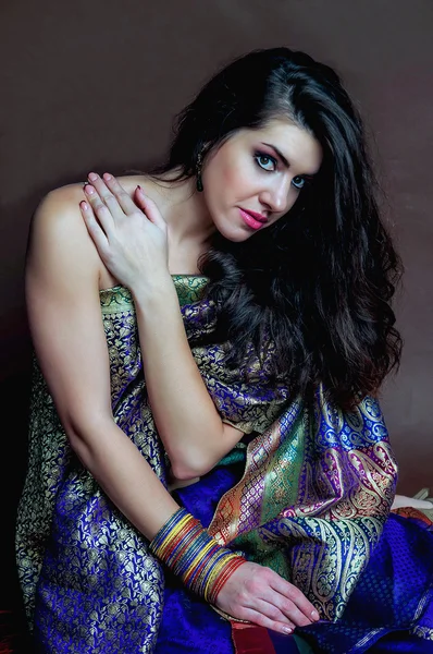 The young dark-haired woman in a bright  Indian sari and colored bracelets sitting and looking thoughtfully touching shoulder hand. Indian style.