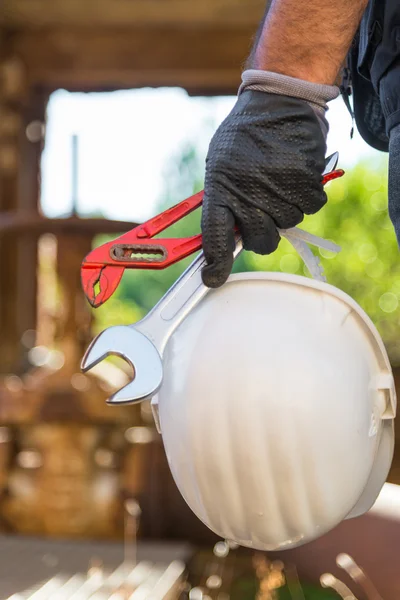 Hands with work gloves holding a safety helmet, a wrench and a red key pipe clamps