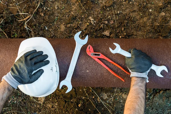 Hands with work gloves holding a safety helmet, a wrench and a red key pipe clamps