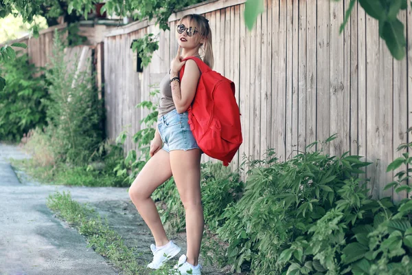 A girl wearing glasses, denim shorts, gray shirt with backpack.