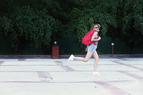 A running girl wearing glasses, denim shorts, gray shirt with backpack.