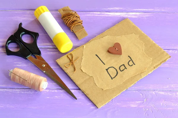 Hand greeting card with message I love dad. Homemade design fathers day card. Happy birthday card for dad. Scissors, glue, cord, thread, needle. Craft inspirations for kids. Lilac wooden background