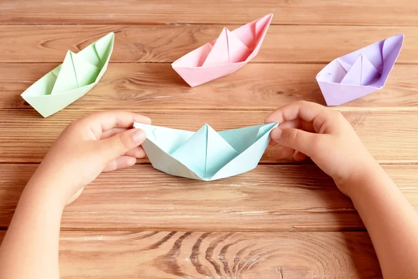 Child holds the origami ship in his hands. Colorful ships origami paper folding on a wooden table