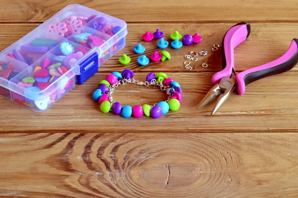 Colorful button charm bracelet, a box of different buttons, pliers, metal connecting rings on wooden table. Using buttons for original fun project. Easy make a bracelet. Hand jewelry
