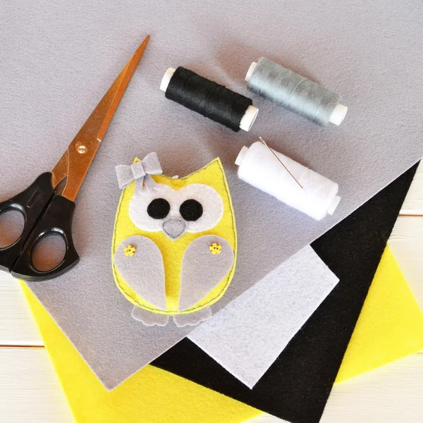 Cute felt owl toy decorated with bow and buttons. Stuffed animal owl on wooden background. Home decor plush toy. Felt sheets, scissors, thread, needle, pins. Top view