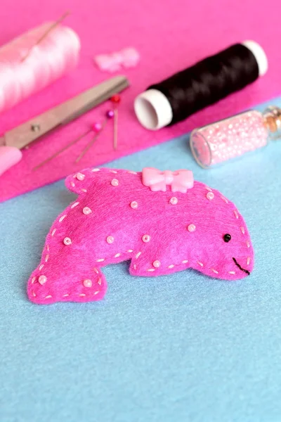 Pink felt dolphin toy on a blue background. Scissors, thread, needles, pins, beads, button. Funny sewing and craft activity for children
