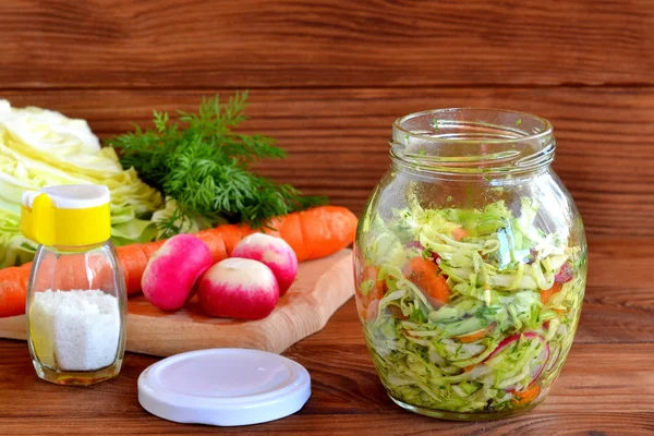 Vegetable mixed salad in a jar. Salad prepared of radish, carrots, cabbage, olive oil, salt and dill. Green dill, fresh carrot, cabbage, radishes on a wooden cutting board. Tasty vitamin food