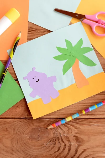 Fun summer card with a hippopotamus and a palm tree on a wooden table. Preschool and kindergarten crafts. Materials for kids art. Paper and glue crafts activity for children. Early child development