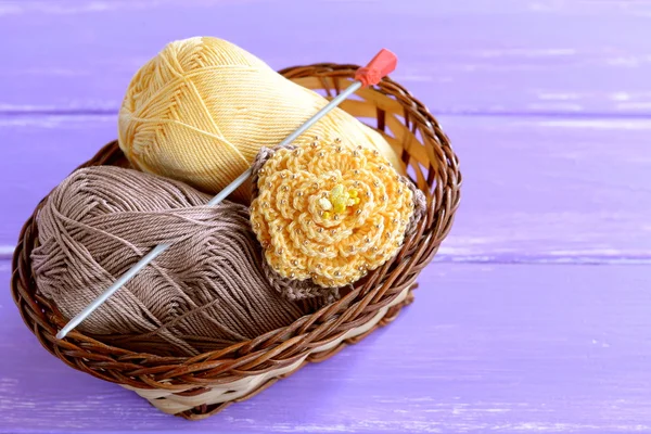 Yellow crochet flower with brown leaves and beads, hook, cotton yarn in wicker basket on lilac wooden background. Easy crochet flower ornament photo. Crafts for children women, beginners