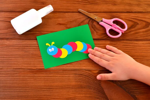 Child holds a paper application. Children paper collage - colored caterpillar on green sheet, glue, scissors. Brown wooden background