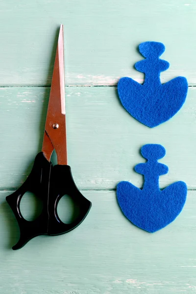Two elements in the shape of an anchor cut from blue felt. Scissors on wooden background. How to sew a fabric toy for children. Hand crafts. Step. Closeup. Top view
