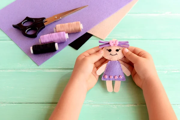 Child holds a small doll in his hands. Doll is sewn from felt. Scissors, thread and felt set on a wooden table. Cute fabric art and craft idea for kids
