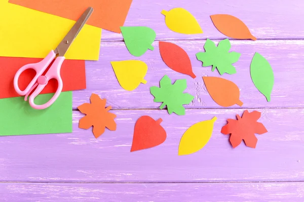 Cut yellow, green, red and orange paper leaves, scissors, colored paper sheets on lilac wooden background. Easy autumn crafts for kids in kindergarten at home. Closeup