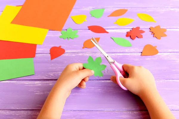 Child cuts out colored paper leaf. Child holds scissors and a green leaf in his hands. Colored paper set, colorful leaves on a wooden table. Autumn kids crafts idea