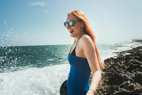 Young woman with red hair and sun glasses standing on sea coast  playing  water spray