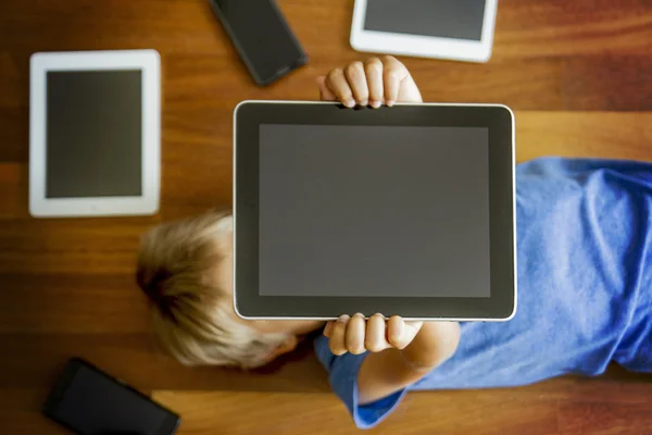 Little boy holding tablet overhead lying on wooden floor. Digital tablets, mobile phones, laptop all around. Top view. Education, learning, technology concept