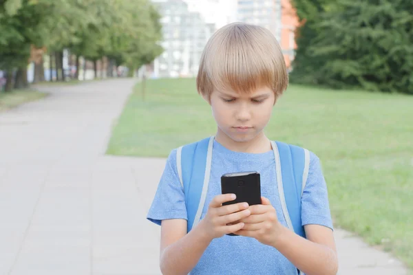 Boy with mobile phone in the street. Child looks at the screen, use apps, plays, writes or reads message. City background. School, people, technology, leisure concept