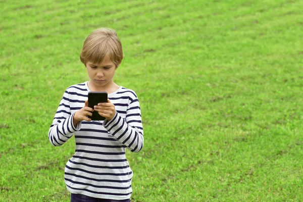 Boy with mobile phone outdoor. Child looking at the screen, playing, using apps. Green grass background. People, technology, leisure concept