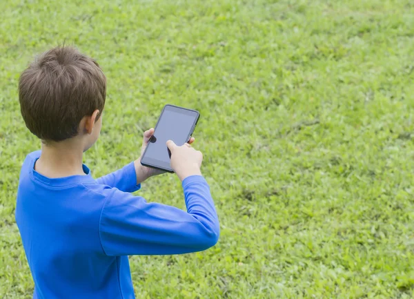 Boy holding mobile phone on green grass lawn with copy space for text. Back view. Technology people connection concept