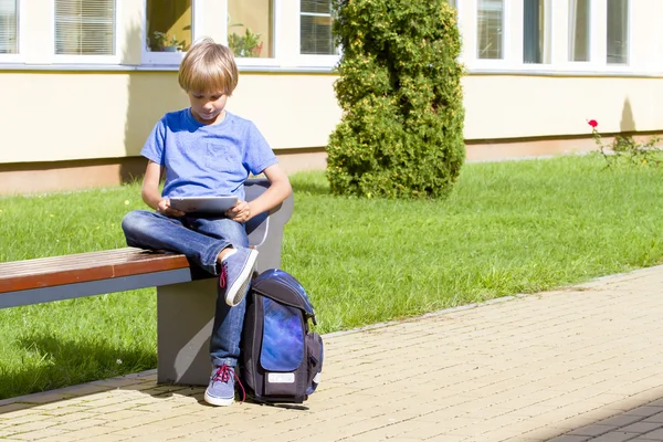 Schoolboy with tablet PC sitting on the bench near school. Outdoor. Education, technology, people concept