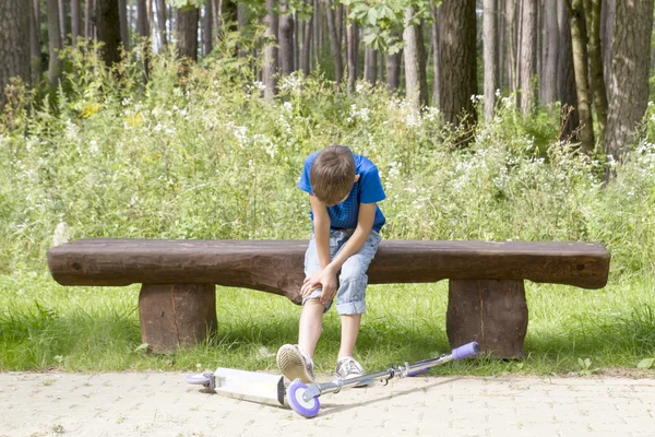 Boy sitting on the wooden bench in the park. Child fell while riding his scooter and hurt leg